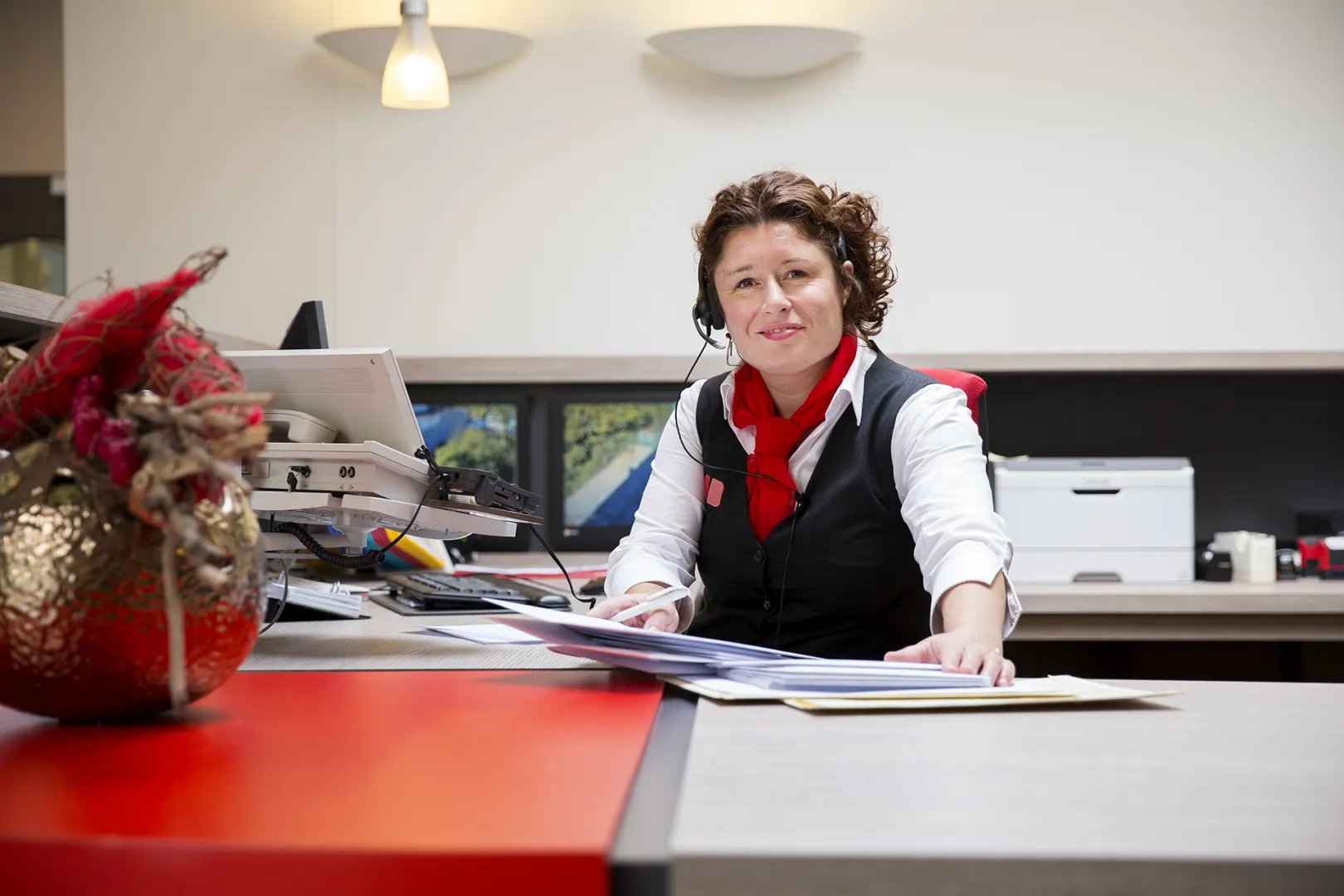 Mevrouw zit achter de balie van de receptie en kijkt naar de camera met een boek op haar bureau.