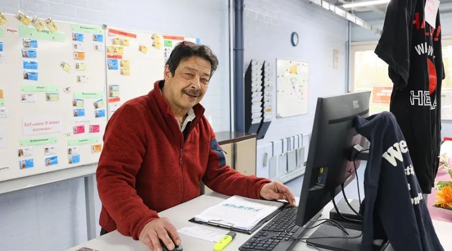 man kijkt lachend in de camera terwijl hij achter een stabureau staat, achter een computer